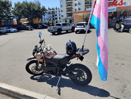 A photo of a Zero DS electric motorcycle covered in queer stickers with a full sized transgender pride flag on a pole mounted securely to the back tail. It's parked in front of Dick's Drive-In.
