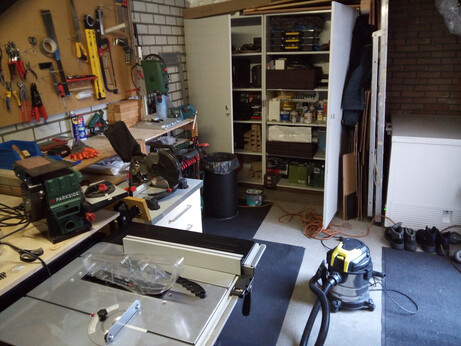 An overview picture of the garage, taken at an angle; everything looks very tightly packed with no gap left unused, except for a large open working space in the middle with rubber mats on the floor. Visible are the table saw in front, a large two-door storage cabinet with shelves in the back (next to a large chest freezer), and some improvised workbenches on the left with a drawer cabinet half slid under one of them. On the drawer cabinet is sitting a miter saw, the workbench contains various assorted items as well as a drill press, and a sanding station is also visible. A hole board is mounted above the workbenches, with various tools hanging and sitting on hooks. Between the chest freezer and the storage cabinets are squeezed a ladder and some cardboard and coats.