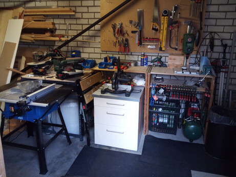 The same workshop from a different angle, pointing at the workbenches this time. Now you can see various items stored under the workbenches; a roll of bubble wrap, a crate of waste, as well as a tank compressor. You can also see one of those foldable workbenches squeezed between a stationary workbench and the drawer cabinet; it is used for outdoor work. A row of clamps is visible, clamped to the under-shelf of one of the stationary workbenches.