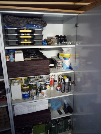 A close-up of the storage cabinet in the back; it contains several parts boxes with handles stacked on top of each other, as well as a stack of carpet tiles, a bag of wallpaper rolls, various covering sheets, a lot of cans of (spray) paint, glue, and so on, and assorted bits like wheels you can screw under something, and some boxes in the bottom containing things that still need to be installed.
