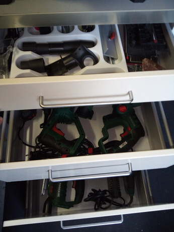 A close-up of the drawer cabinet, with all drawers opened in a staggered manner to show the contents; the bottom two drawers contain various green-and-red Parkside powertools, whereas the top contains a cutlery tray that's actually filled with spare bits and add-on tools for the powertools, as well as a few items next to the cutlery tray (which is undersized for the drawer).