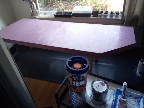 The plant table on top of some protective floor covering, painted in lavender purple but with the legs still being unpainted wood. there is a corner cut out of the tabletop. If you look carefully, you can see that the tabletop is made up of several separate boards that have been glued together. The paint is still drying, looking uneven, and in front of the table is a black metal cart with a can of paint, a brush sitting on a painting tray, and a tall cylinder marked 'brush saver' with a brush stored inside of it.