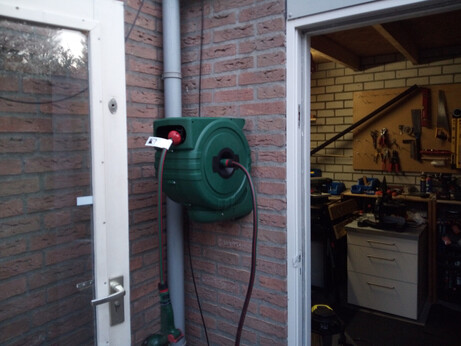 Green box with auto-retractable garden hose coming out of it, mounted on the outside wall of the garage, in a corner. There's a spray head on the end of the hose. On one side of the box you can see the back door of the house being open, on the other side you can see inside the garage, showing the tool board and the work surfaces.