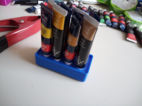 A blue 2x1 (height 3) Gridfinity box, but with holes that are sized exactly to hold small 12ml acrylic/oil paint tubes. The tubes contain a variety of yellow/brown-ish colors. A row of unstored paint tubes is scattered behind the storage bin, and there's an unidentified clamp on the left.