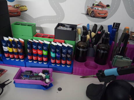 24 paint tubes, arranged cap-down by color into three 3D-printed holders, alongside many other 3D-printed Gridfinity bins holding, among other things, IPA and vinegar bottles, cleaning swabs, pens and markers, a ruler, tweezers, a screwdriver, prototype boards, and assorted electronic components. In the bottom right corner you can see my Pinecil soldering iron sitting on a stand that also holds a tip cleaning thing. In front of the Gridfinity arrangement are two stacked spout bins filled with various electrical components, not part of the storage grid.