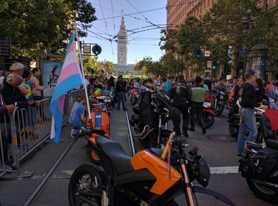 An orange 2013 Zero DS Electric motorcycle with a flag pole mounted off the back flying the trans pride flag at 2016 SF Pride. 
