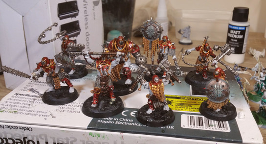 A half-finished group of Iron Golem miniatures from the tabletop game WarCry. They are grey-skinned humanoids wearing red and gold armour, with bronze and steel weapons, and steel spikes protruding from their armour and limbs.
