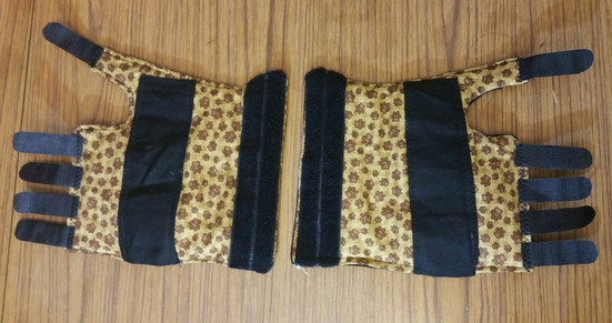 Images show a pair of wristbraces, made from a light brown pawprint fabric, black velcro and black cotton drill for the splint section. This image shows them laid flat, the other two images show the right-hand one being modelled (with splint inserted)