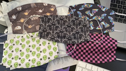 Five fabric face masks. Top left has illustrations of Honduran white bats and big brown bats on it. Top right has a rainbow coloured bat design. Centre has a dark floral pattern with bat silhouettes hidden in it. Bottom left has a pattern of alien heads and stars, matching symbology from Steven Universe. Bottom right has the missing texture pattern from Source engine games. 