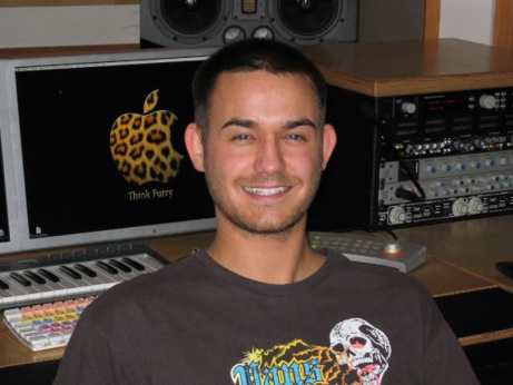 A smiling white guy in front of a music production setup, the wallpaper has an Apple logo with a leopard texture with "Think Furry" below it