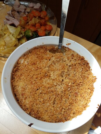 Cooked mac and cheese in a circular baking dish with a spoon. Plate of ham, carrots, cabbage, and brussel sprouts in the background
