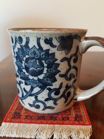 Grey coffee mug with a stylized flower and vines in shades od navy blue sitting on a red coaster with tassels