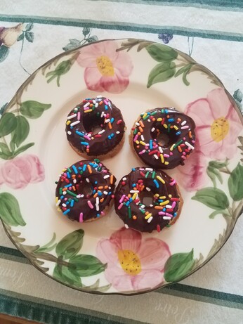 Four mini buttermilk doughtnuts with chocolate glaze qnd rainbow sprinkles on a desert rose plate of a cream plate with pink flowers and green leaves